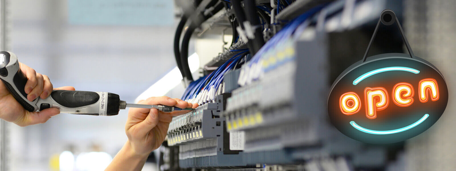Eine Person schraubt mit einem Akkuschrauber an einem Schaltschrank und im Vordergrund hängt ein schwarzes Schild mit Neonlinien und dem neonfarbenen Schriftzug "open"